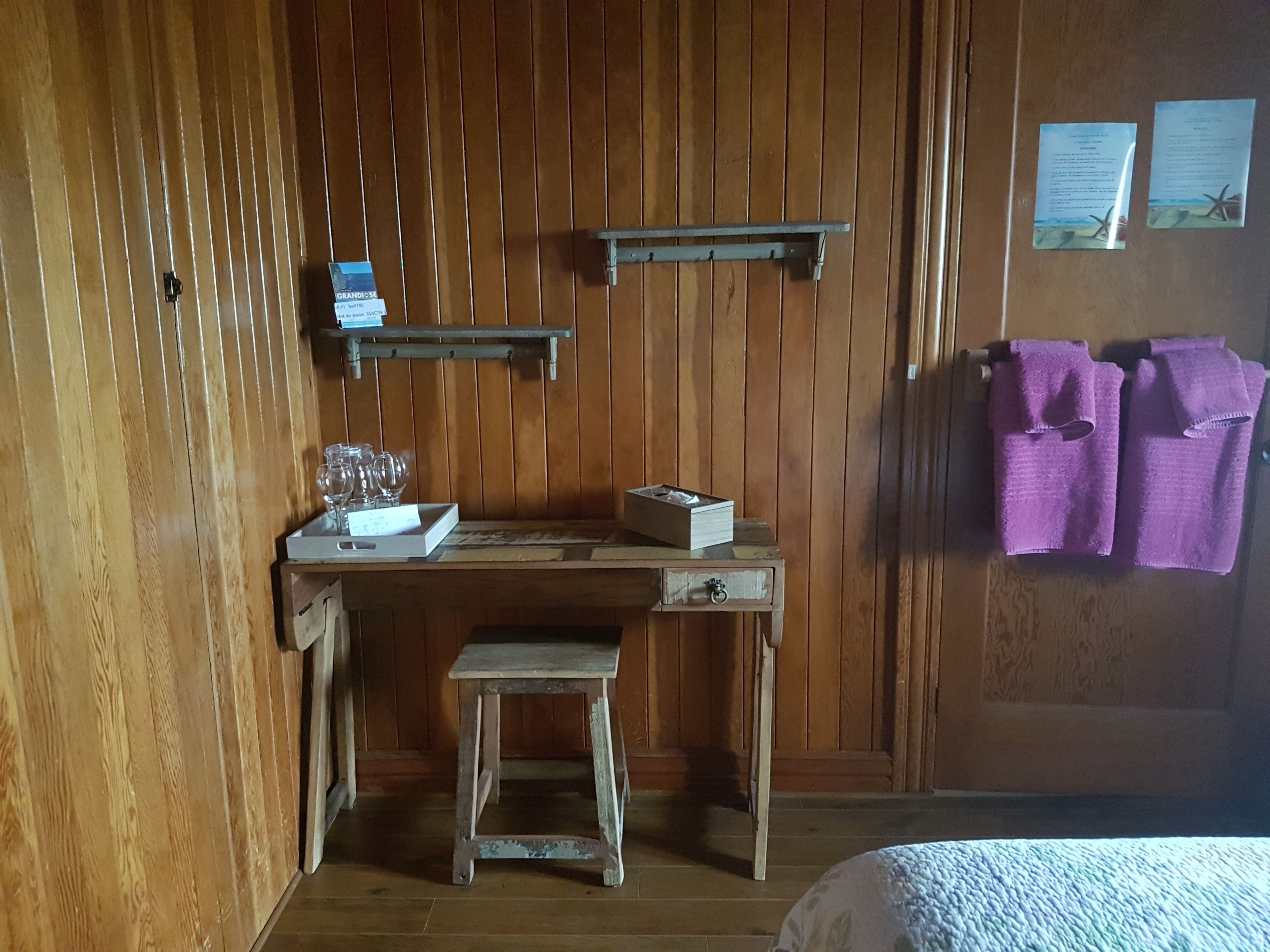 A wooden room with a small desk, a stool, and a bed. There are towels hanging on the wall and a shelf with items on it.