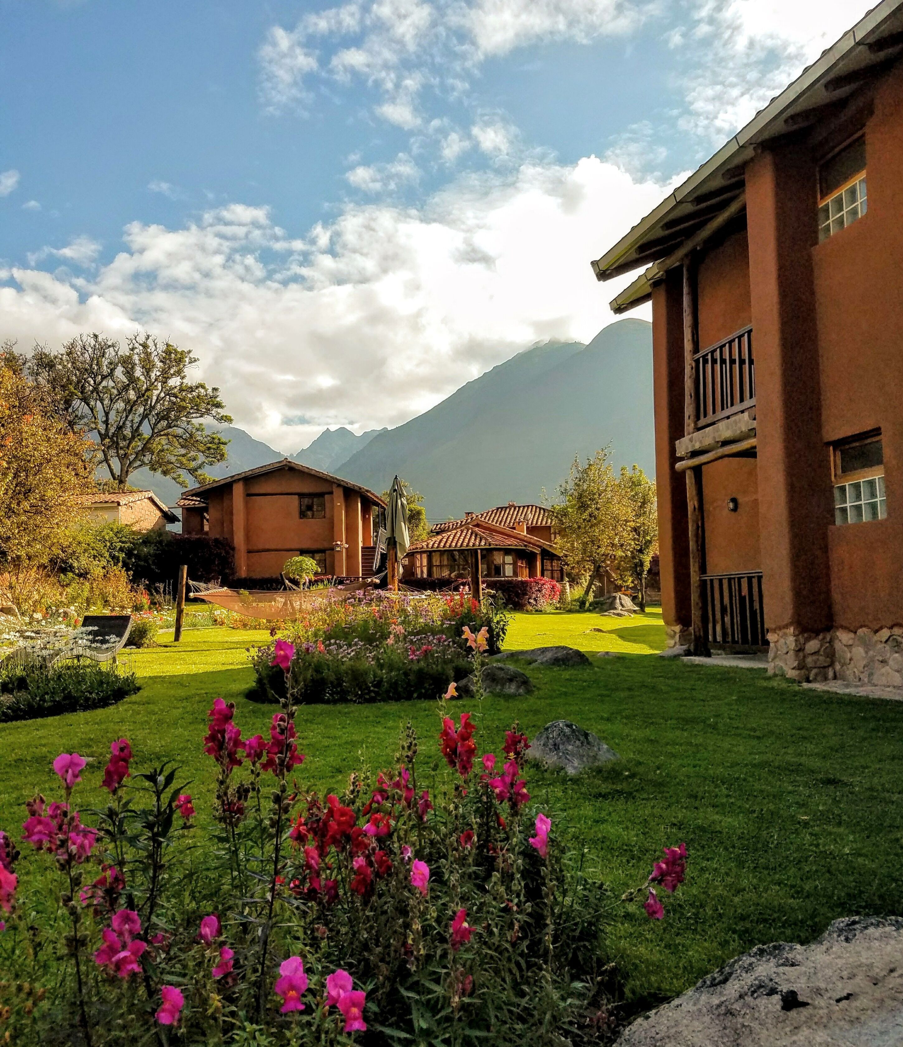 A traditional building with a stone facade and a balcony, surrounded by a well-maintained garden with flowering plants and a clear view of mountains in the background.
