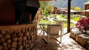 A stone fireplace with a chimney, a glass table, and two chairs in a covered outdoor area with a view of a garden and buildings in the background. - Lizzy Wasi Urubamba (Urubamba)