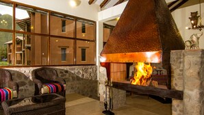 A stone fireplace with a fire, wicker chairs with colorful cushions, and a view of a mountain through large windows. - Lizzy Wasi Urubamba (Urubamba)