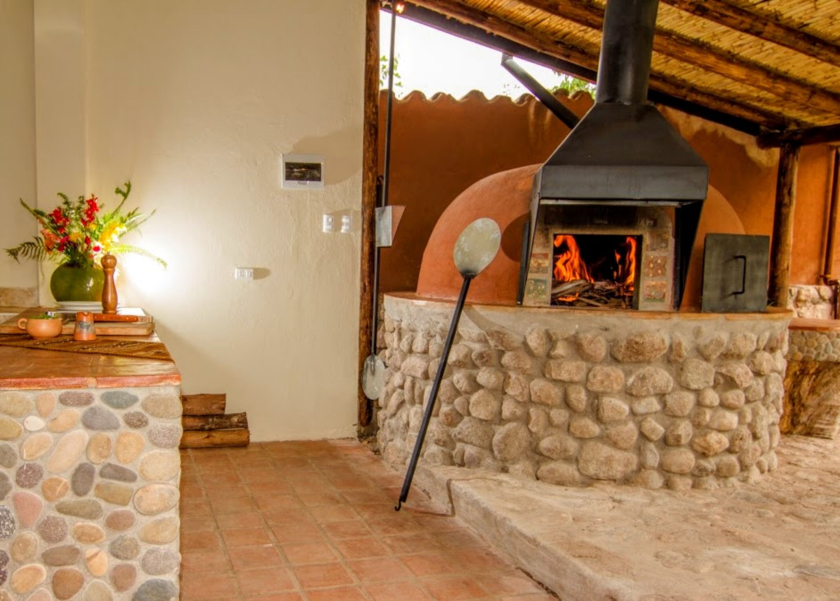 Un patio empedrado con un horno rústico al aire libre, un muro de piedra y una pequeña mesa con un florero.