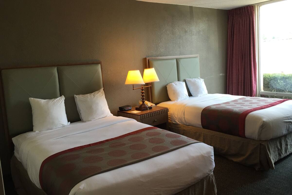 A hotel room with two beds, each with white linens and a red patterned throw. There are two bedside tables with lamps, and a window with red curtains.