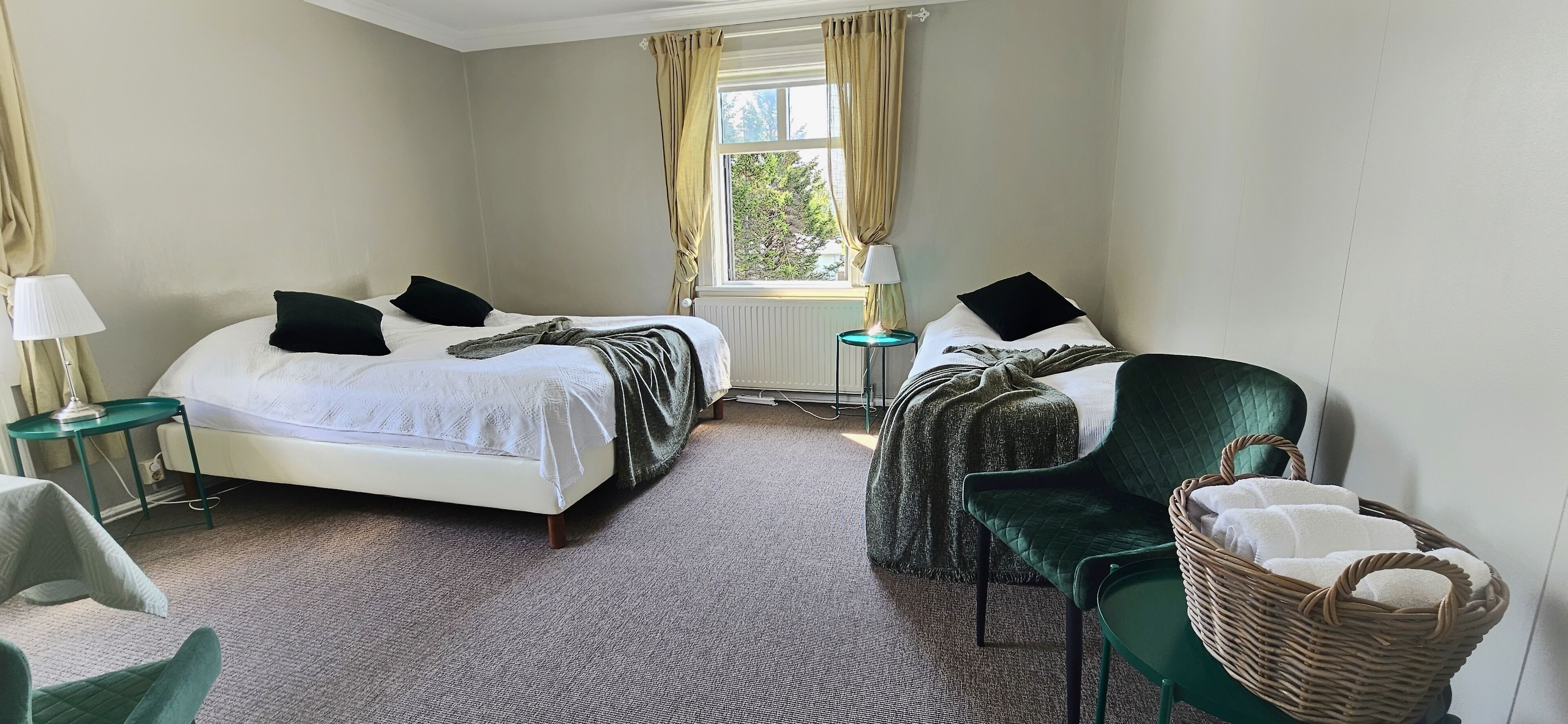 A hotel room with two beds, a green armchair, a wicker chair, and a small table with a lamp.