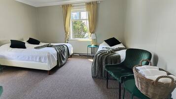 A hotel room with two beds, a green armchair, a wicker chair, and a small table with a lamp.