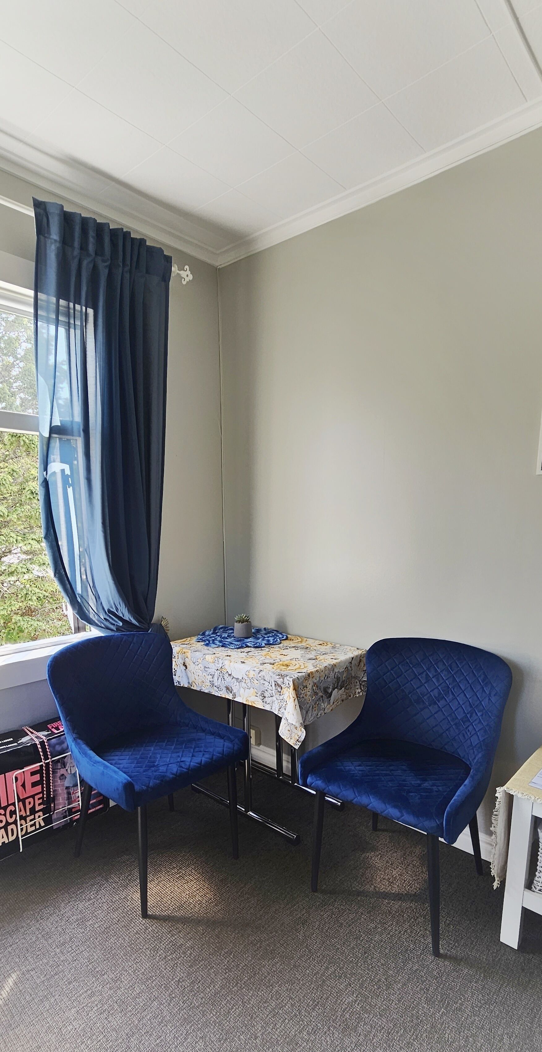 A corner room with two blue chairs, a small table with a floral tablecloth, and a window with blue curtains.