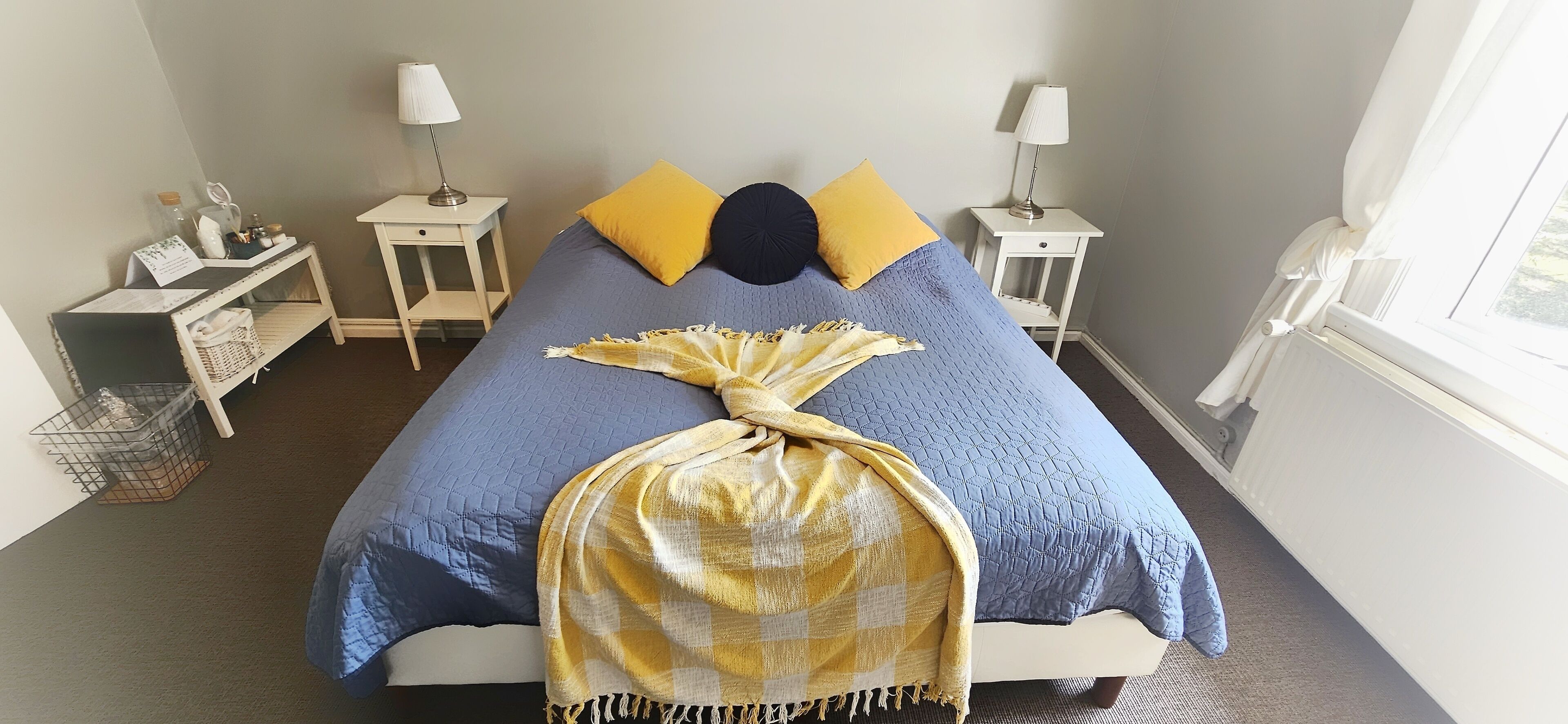 A neatly made bed with a blue quilt and two pillows, one yellow and one dark blue. There are two bedside tables with lamps and a small shelf with personal items.