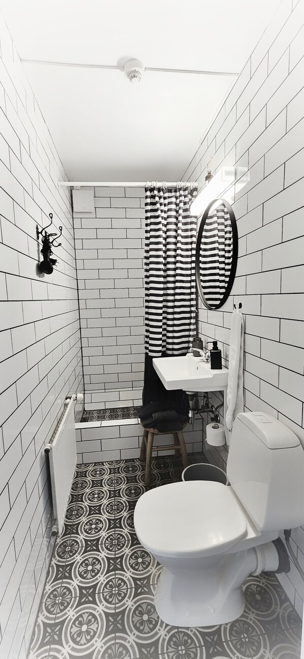 A compact bathroom with white tiled walls, a black and white striped shower curtain, a white sink, and a toilet. - Árból Guesthouse (Husavik)