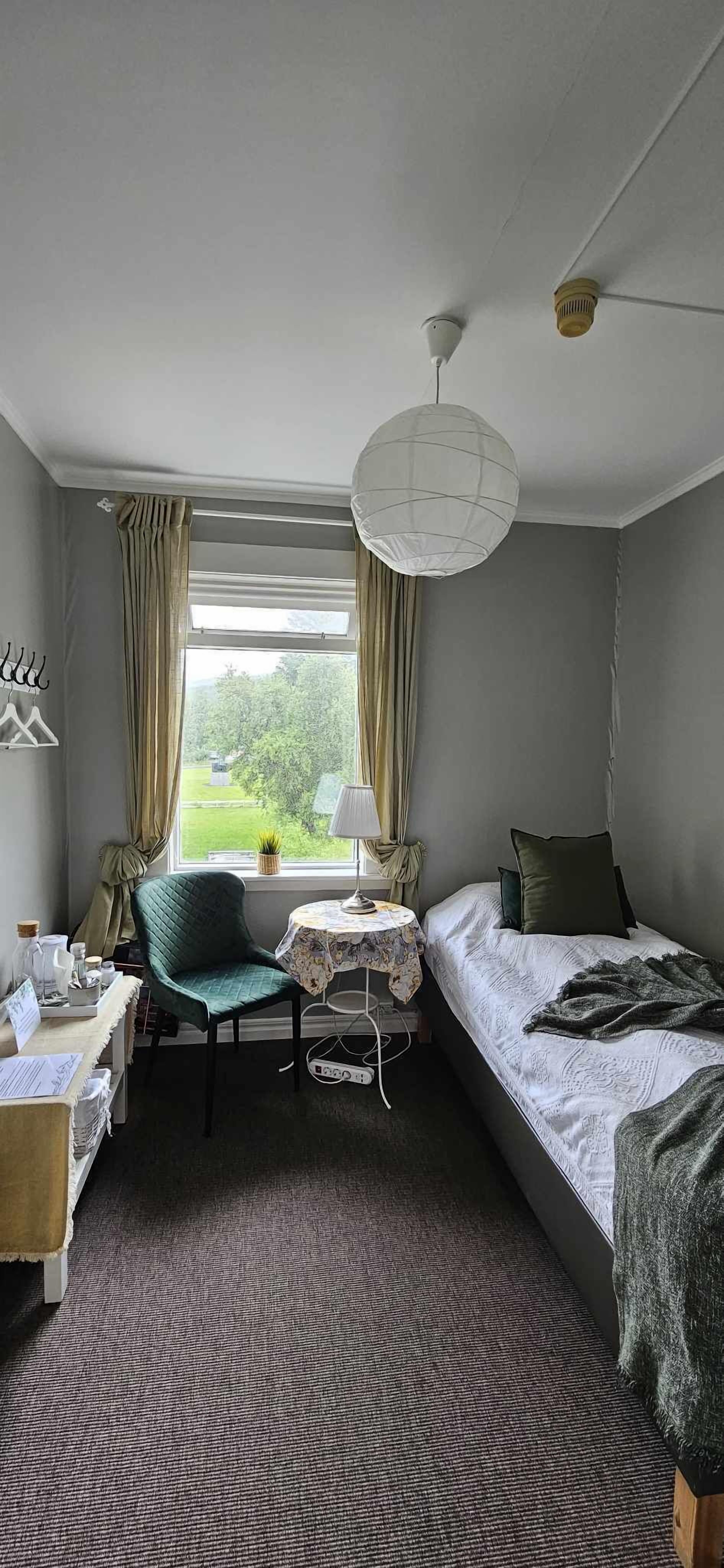 A bedroom with a bed, a desk, a chair, a lamp, and a window with curtains.