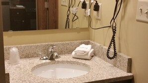 Un baño con lavamanos de encimera de granito, un espejo, dos secadores de pelo montados en la pared y un cubo de basura.
