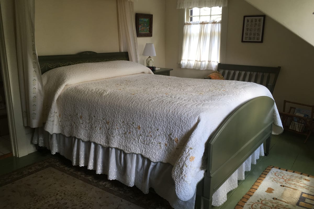 A bedroom with a four-poster bed, a nightstand, a lamp, a window with curtains, and a framed picture on the wall.