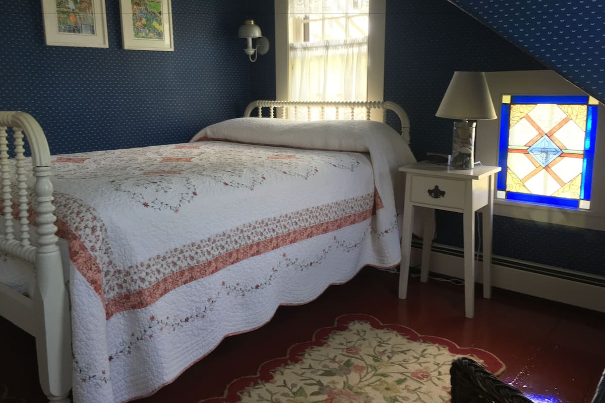 A bedroom with a bed, a nightstand, a lamp, a window with a stained glass design, and two framed pictures on the wall.