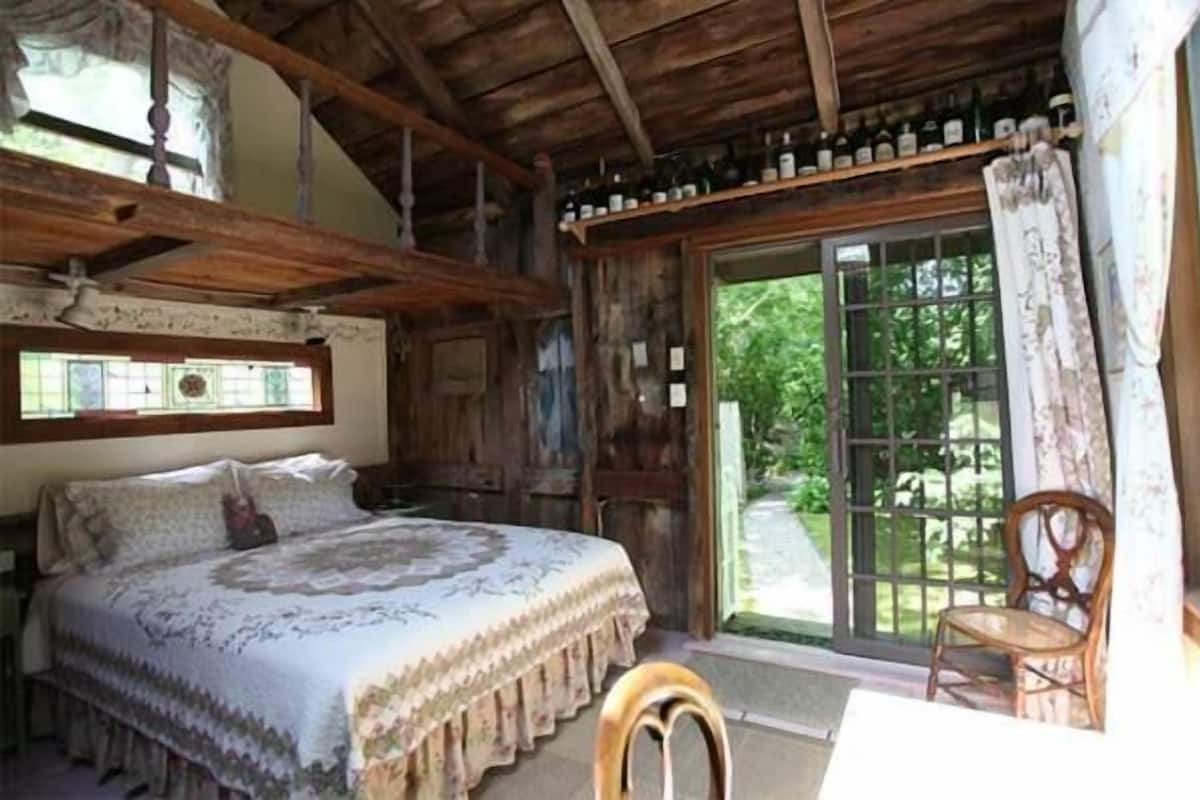 A rustic wooden room with a large bed, a chair, and a window with a view of greenery.