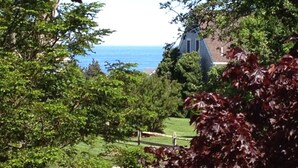 En vy över en fastighet vid kusten med ett stort hus, frodig grönska och fri utsikt över havet.