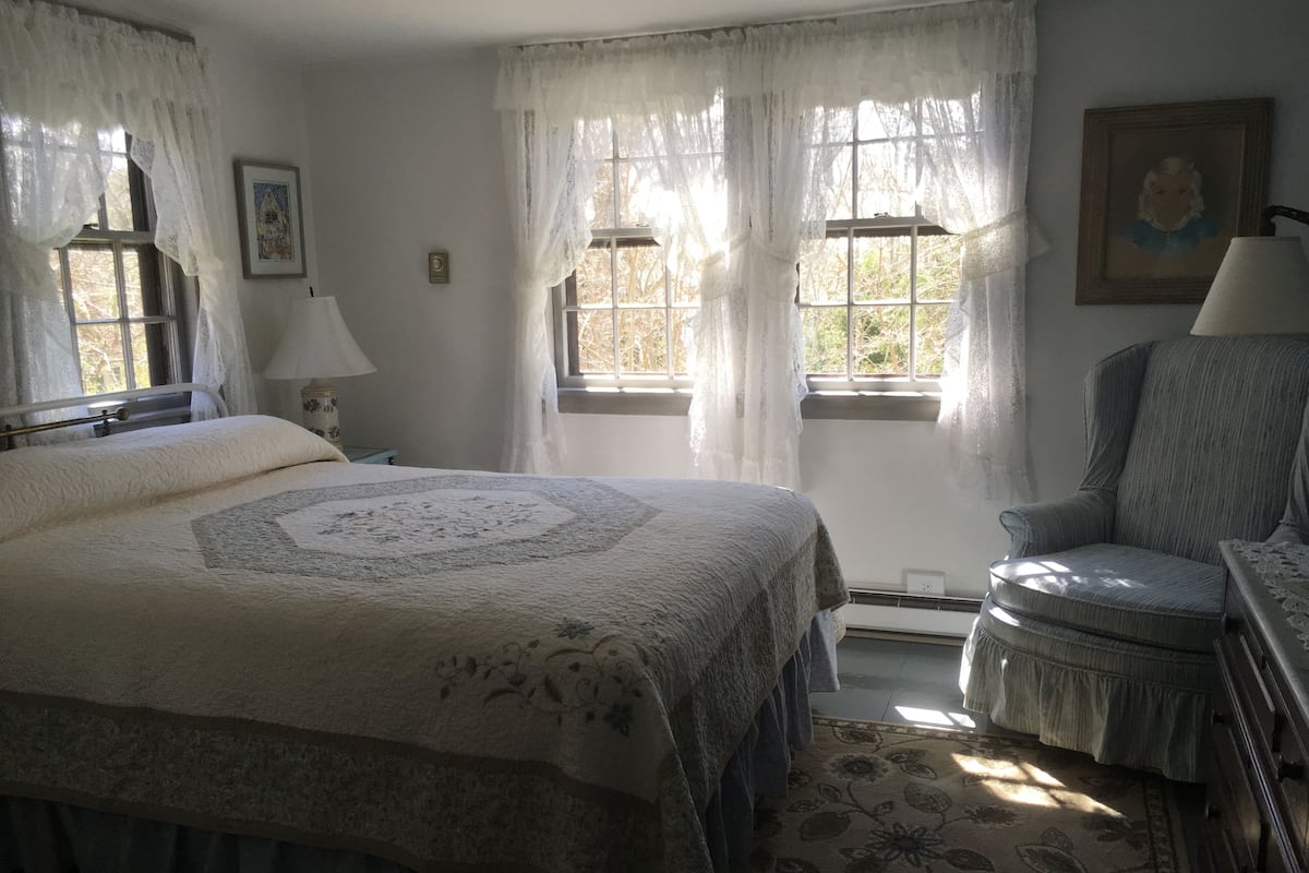 A bedroom with a large bed, two windows with sheer curtains, a chair, a bedside table, and a lamp.