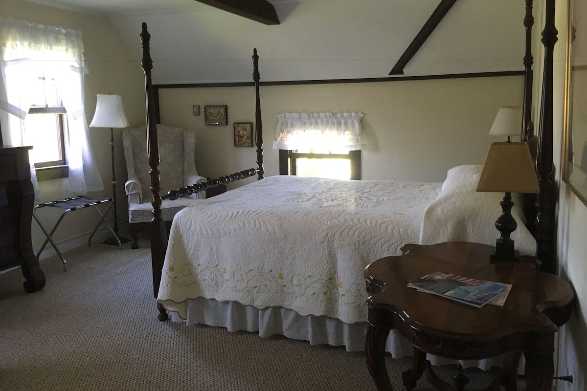 A four-poster bed with a white bedspread, a wooden nightstand with a lamp, a chair, and a small table with a magazine.