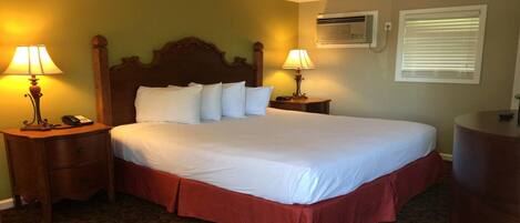 A hotel room with a large bed, two bedside tables with lamps, a bedside phone, a wall-mounted air conditioner, and a window with blinds.