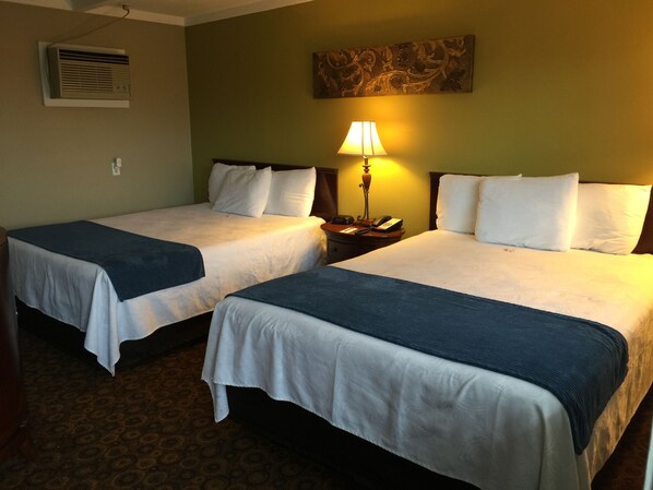 Two single beds with white linens and dark blue throw blankets, a bedside table with a lamp, and a wall-mounted air conditioner.