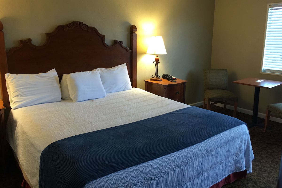 A neatly made bed with white and blue bedding, a wooden headboard, a bedside lamp, a chair, and a small table.