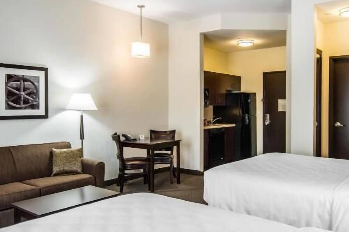 A hotel room with a sofa, a small table, two beds, a desk, a chair, and a kitchenette.