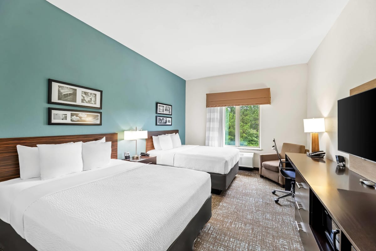 A hotel room with two beds, a desk, a TV, and a window with a view of greenery.
