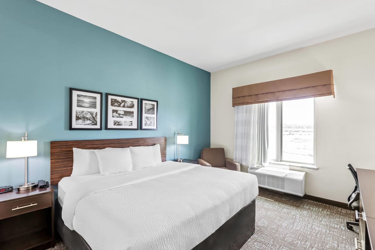A hotel room with a large bed, a nightstand with a lamp, a chair, and a window with curtains.