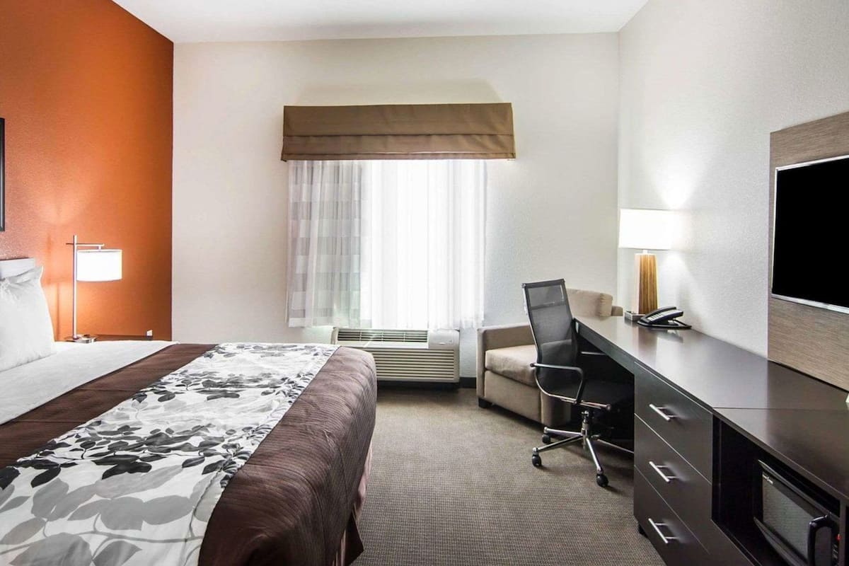 A hotel room with a bed, desk, chair, TV, and window with curtains.