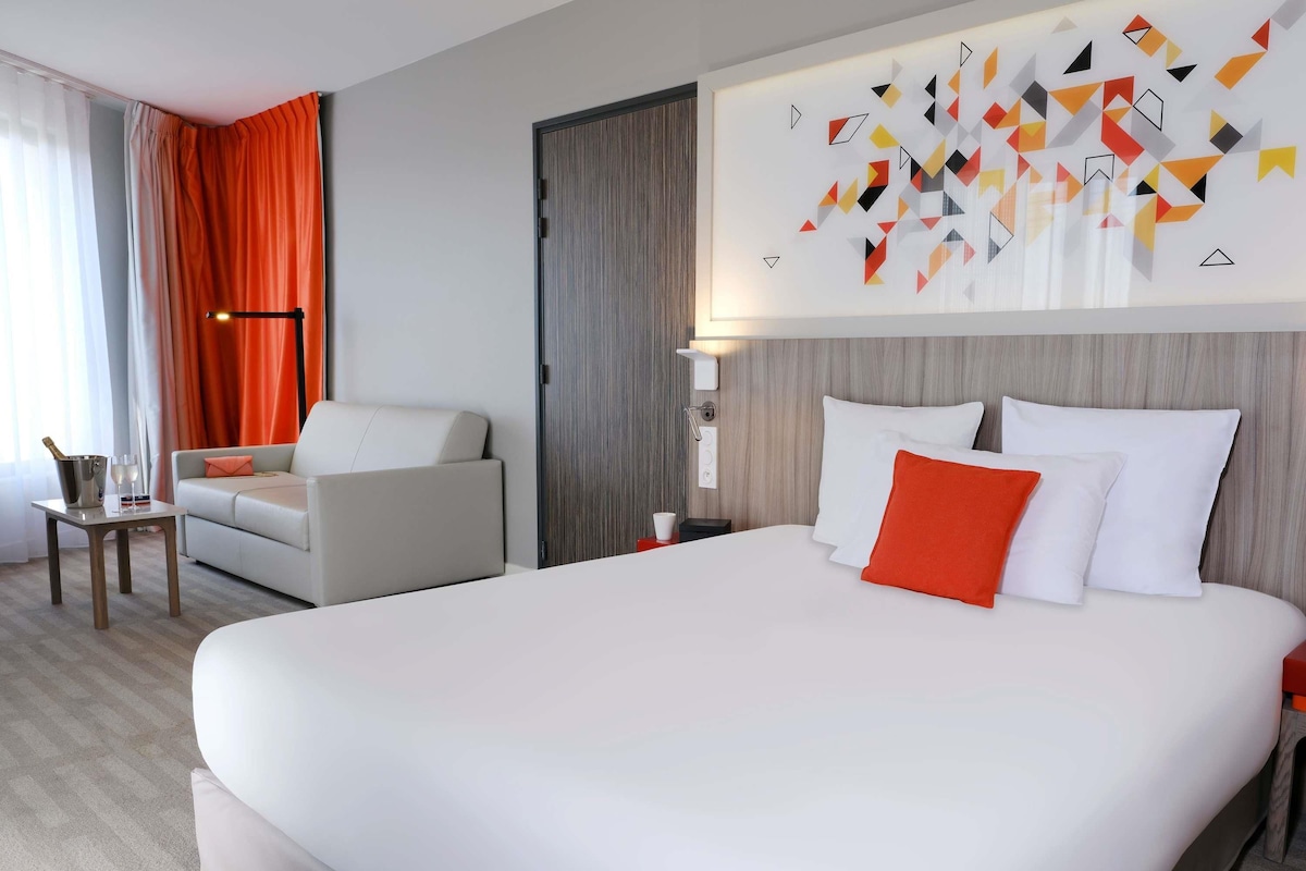 Una habitación de hotel moderna con una cama grande, un sofá, una mesita con una botella y vasos, y una pared decorada con arte geométrico.