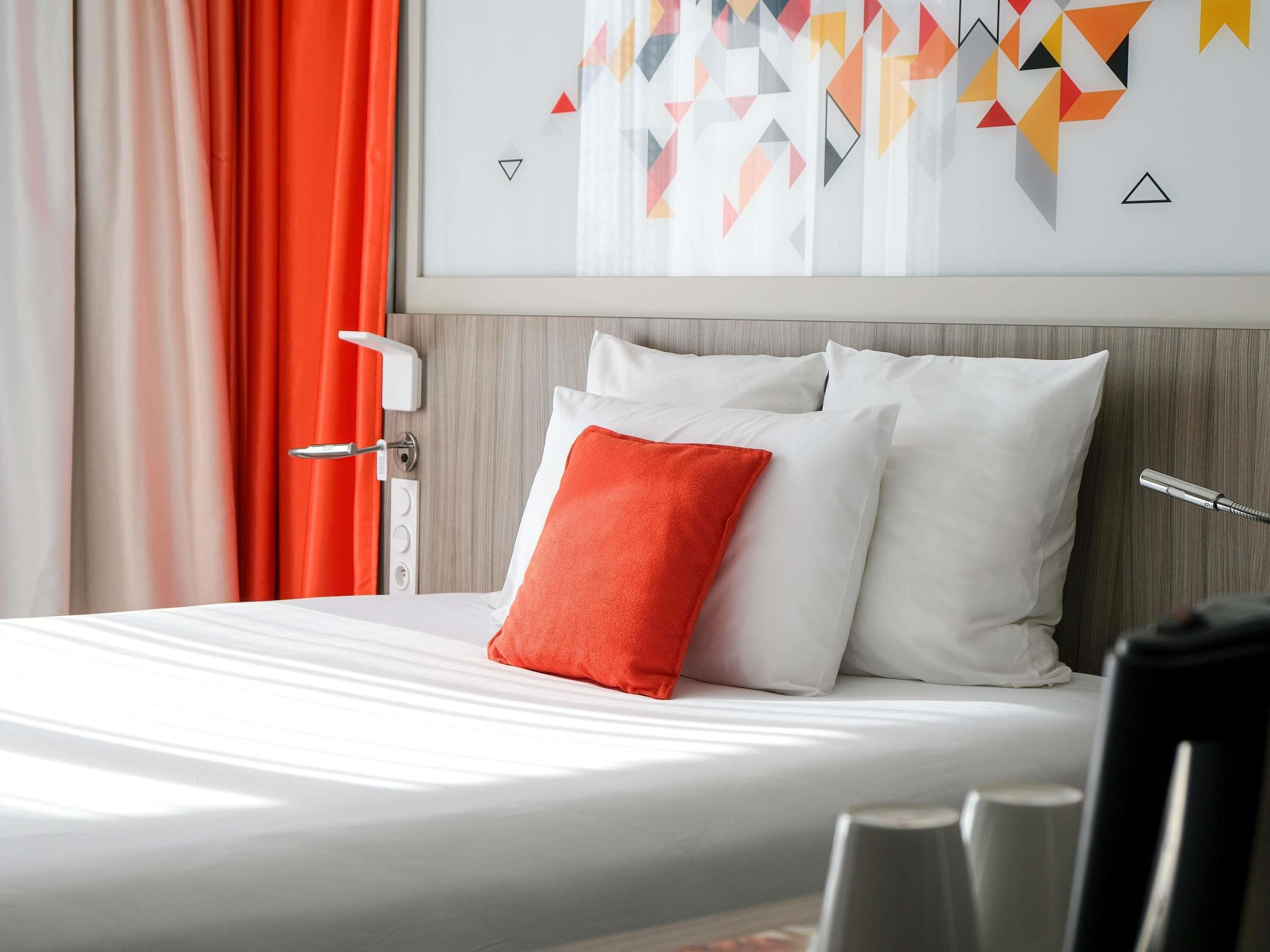 A hotel room with a bed, white pillows, a red pillow, a shower control panel, and a geometric wall art piece.