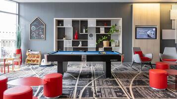 Una mesa de billar en una habitación con alfombra de diseño geométrico, taburetes rojos y una estantería de pared con varios objetos.