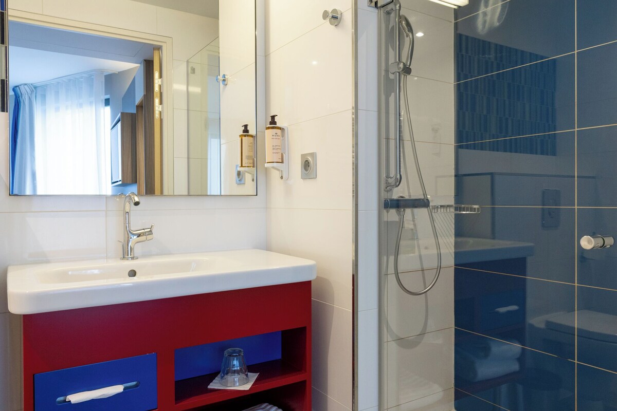 A bathroom with a white sink, red vanity, a mirror, a shower, and a glass-enclosed shower area.