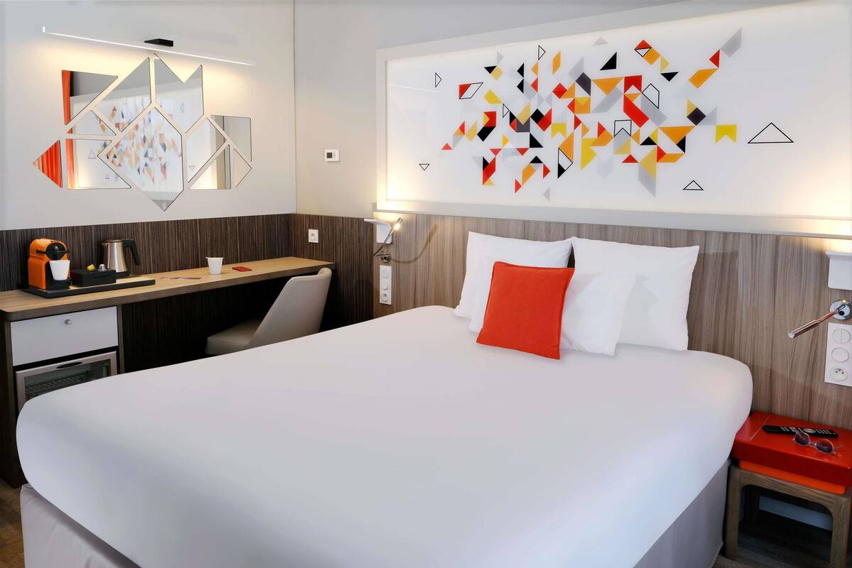 A modern hotel room with a large bed, a desk, and a coffee machine.