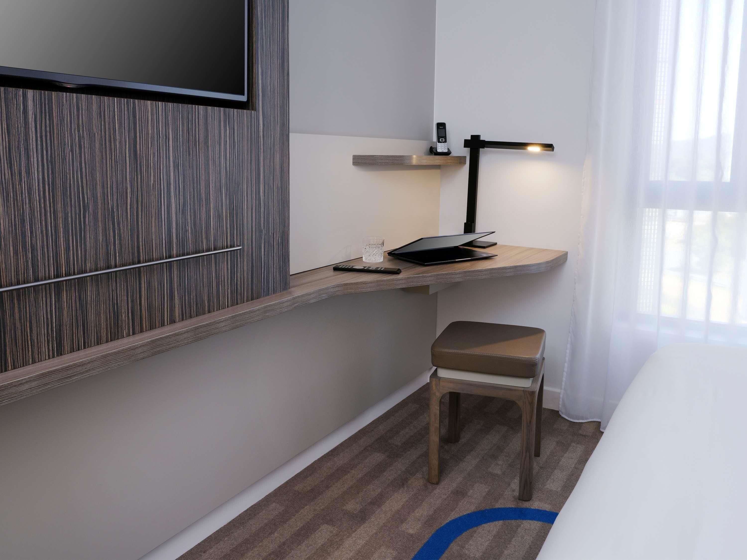 Quarto de hotel pequeno com uma escrivaninha de madeira, um laptop, um telemóvel e uma cadeira.