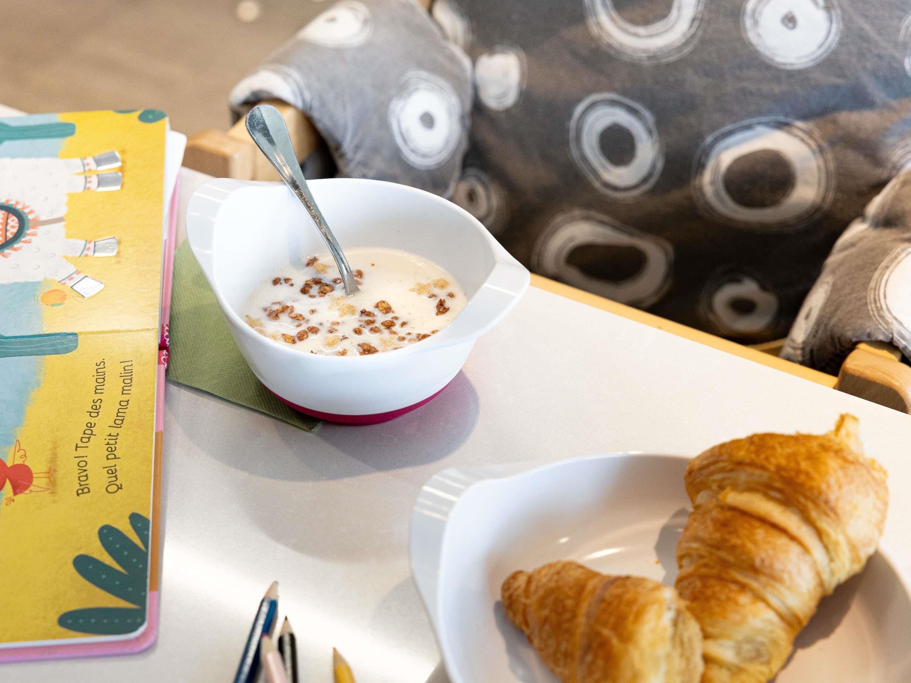 A breakfast setting with a bowl of cereal, a croissant, and a book on a table.
