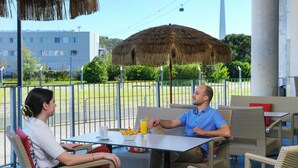 A couple dining at an outdoor cafe with wicker furniture and a thatched umbrella.
