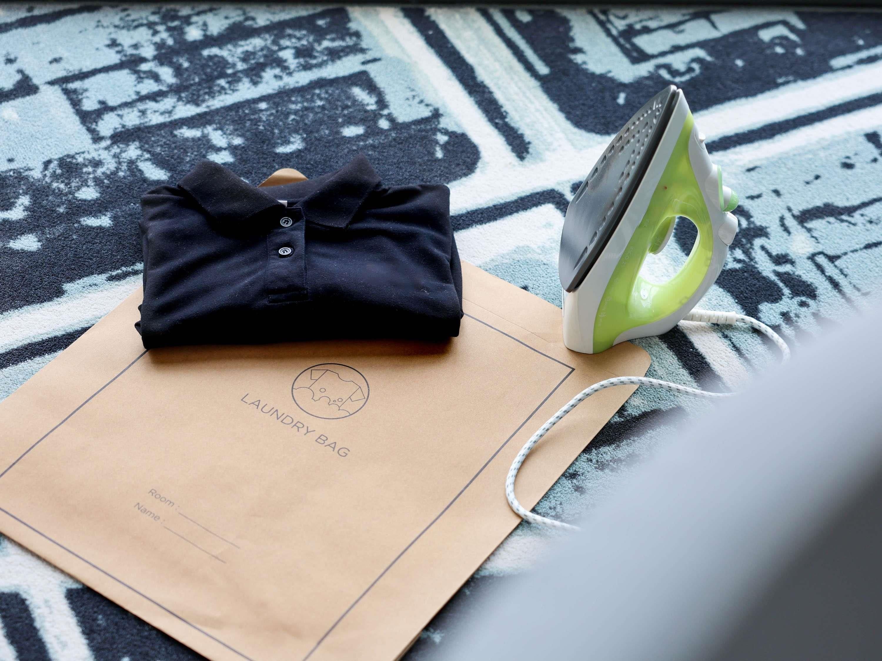 Una camisa azul oscuro bien doblada sobre una bolsa de papel marrón con la inscripción 'LAUNDRY BAG', y un plancha verde y blanca sobre una alfombra estampada.