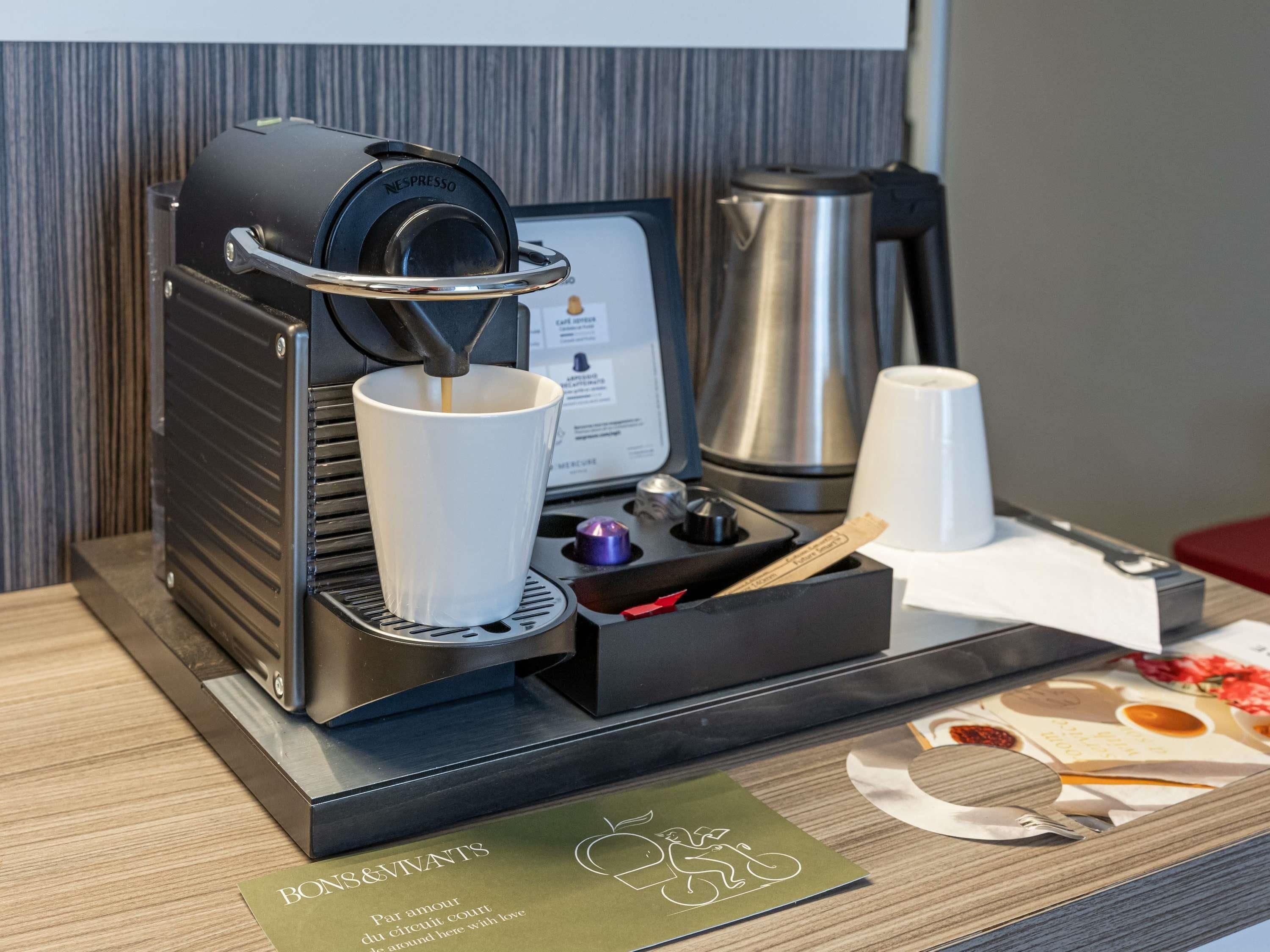 Uma máquina de café Nespresso com um porta-xícaras, um jarro de café de aço inoxidável e um cardápio sobre uma mesa de madeira.