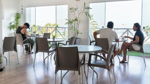 A modern indoor dining area with round tables and chairs, large windows, and a view of the outdoors. - Hostal Los Frayles (Paracas)