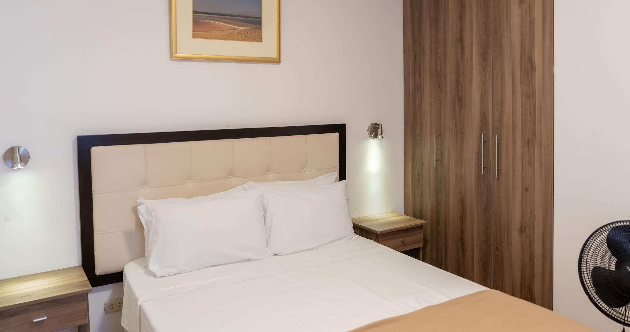 A neatly made bed with a headboard, two bedside tables with lamps, a wooden wardrobe, and a framed picture on the wall.