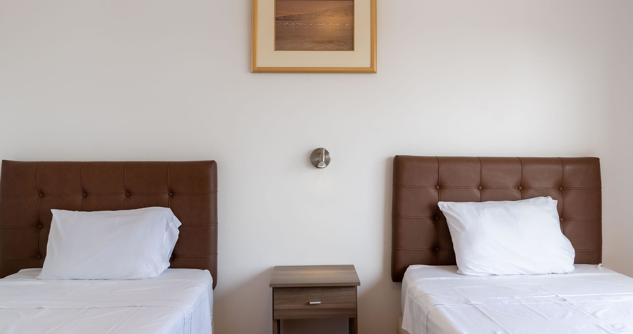 Camera d'albergo con due letti, ciascuno con una testata marrone e biancheria da letto bianca. Tra i letti si trova un comodino in legno, e sopra il muro è appesa una cornice con un'immagine.