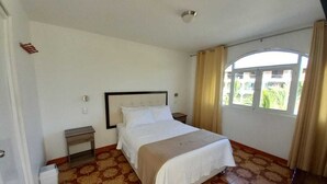 A bedroom with a bed, a nightstand, a small table, a window with curtains, and a patterned floor. - Hostal Los Frayles (Paracas)