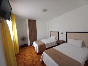 A hotel room with two beds, a fan, a TV, and a window with curtains. - Hostal Los Frayles (Paracas)