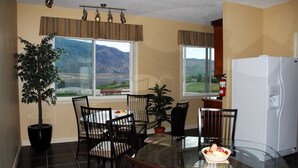 A dining area with a glass table, black chairs, a potted plant, and a view of mountains through large windows. - Ciliegia Villa (Osoyoos)