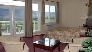 A bedroom with a bed, two armchairs, a glass-top table, and a view of a mountainous landscape through large windows. - Ciliegia Villa (Osoyoos)