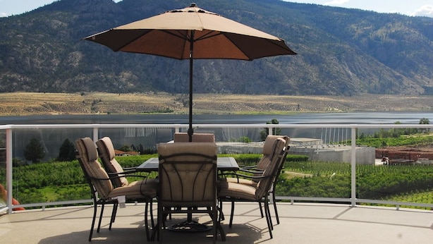 A patio with a table and chairs, an umbrella, and a view of a lake and mountains. - Ciliegia Villa (Osoyoos)