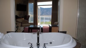 A bathroom with a large bathtub, a television mounted on the wall, and a view of a lake and mountains through the window. - Ciliegia Villa (Osoyoos)