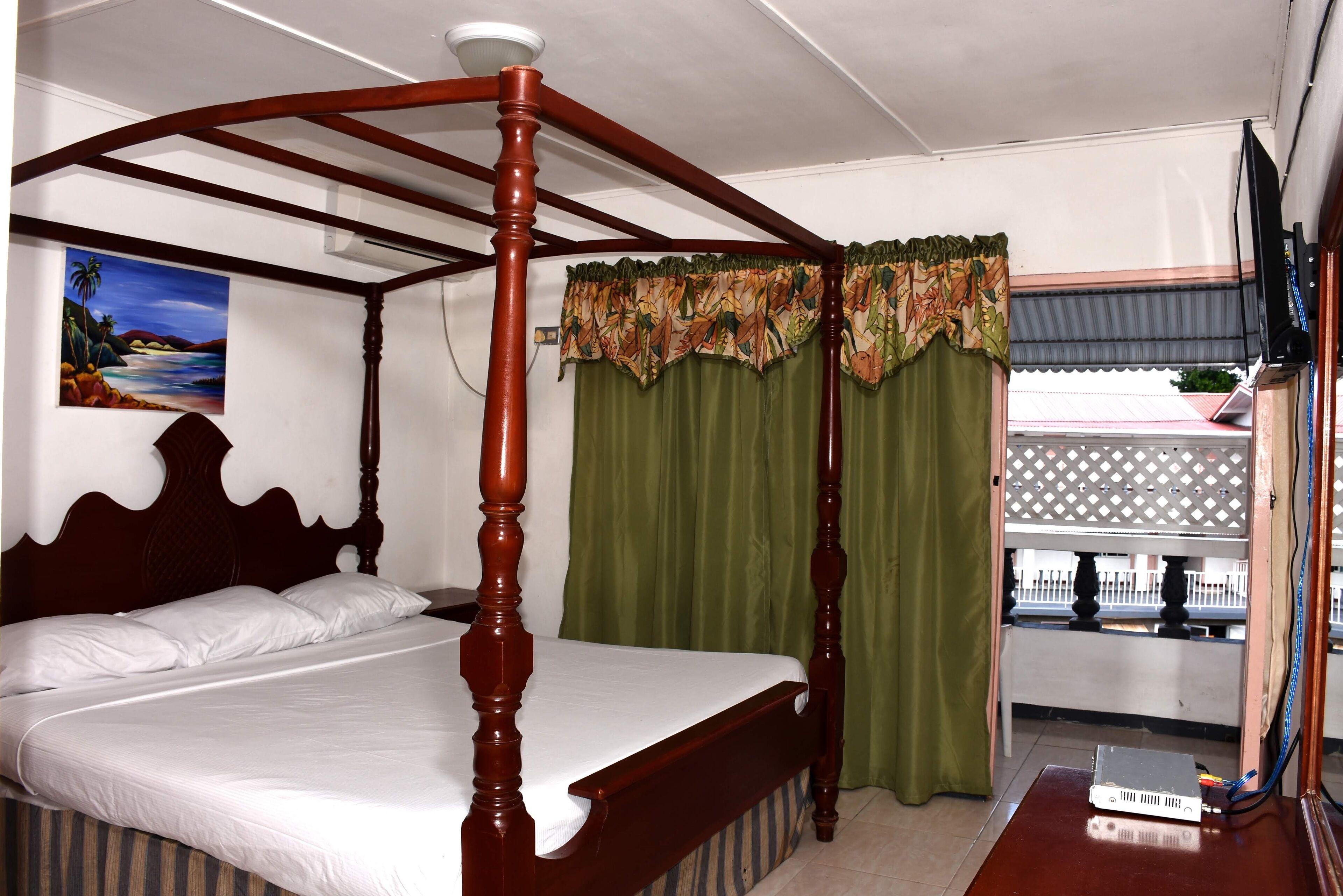 A four-poster bed with a wooden frame and white bedding, a painting of a tropical scene on the wall, and a green curtain.