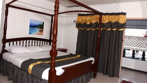 A four-poster bed with a dark wooden frame, a white mattress, and a black and gold bedspread. There is a painting of a beach scene on the wall above the bed. - Marine View Hotel (Ocho Rios)