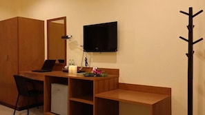 A compact room with a wooden desk, a chair, a small refrigerator, a mirror, a television, and a coat rack.