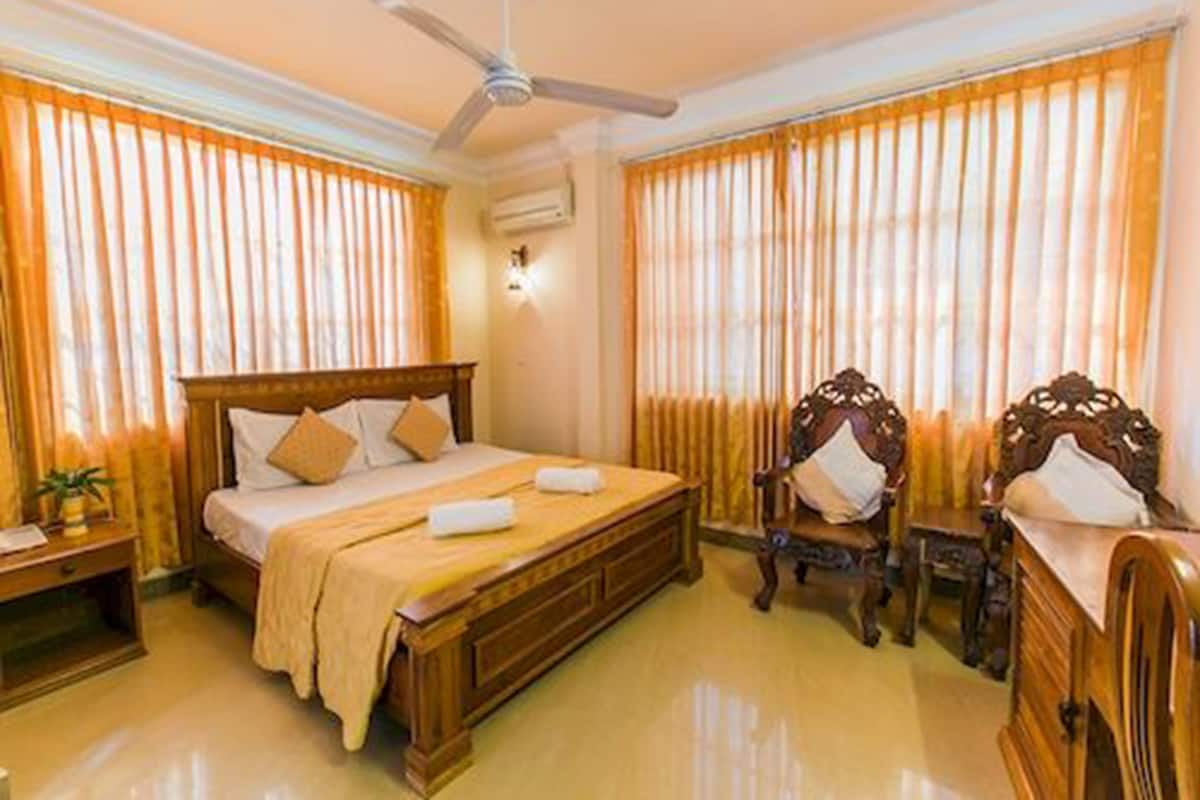 Une chambre d’hôtel avec un lit en bois, un ventilateur de plafond, une table de chevet et deux fauteuils rembourrés.