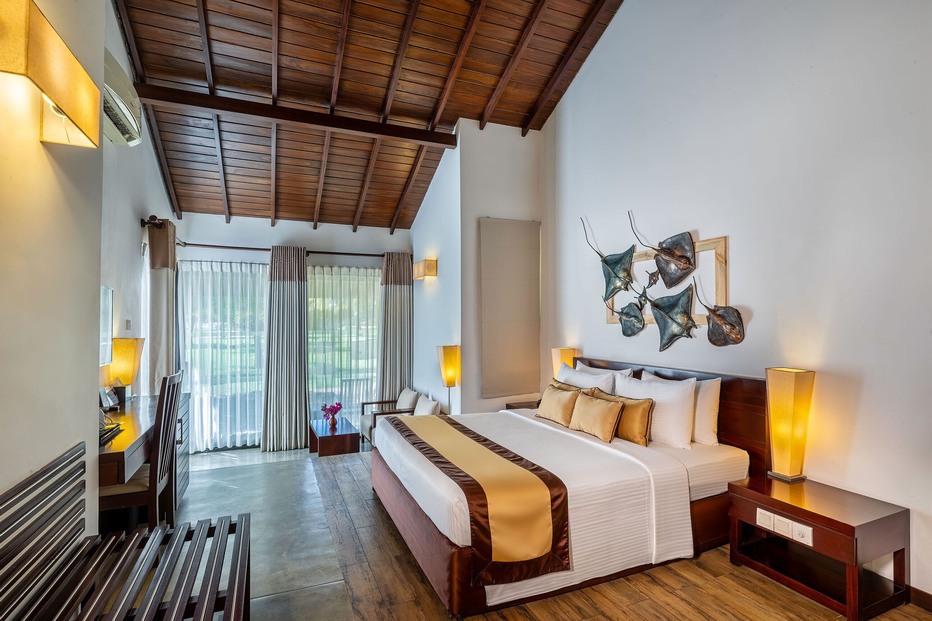 A hotel room with a large bed, wooden flooring, a desk with a lamp, and a view of the outdoors through the window.